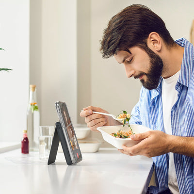 Photo of a man eating while on a video call with his phone attached to a StandyBank with kickstand extended in front of him#color_black